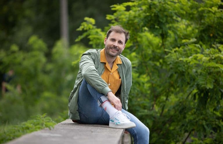 Porträt von Lukas Berger. Lukas hat braune kurze Haare, sitzt auf einer Mauer in einem Park. Er hat weiße Turnschuhe, Jeans, eine grüne Jacke und ein gelbes Hemd an.
