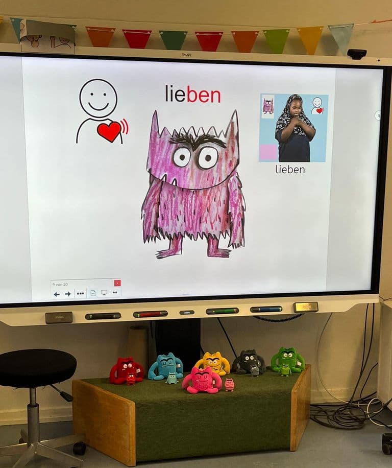 Blick in einen Klassenraum. Auf einem Smartboard ist das METACOM-Symbol für "lieben", und ein rosa Gefühlemonster zu sehen. Am Rand ist eine Karte, mit vier Elementen: Gefühlemonster, die Farbe rosa, ein Screenshot aus der EiS-App auf dem ein Mädchen die Gebärde für "lieben"zeigt und das METACOM-Symbol für "lieben". Unter dem Smartboard sieht man 6 genähte Gefühlemonster.